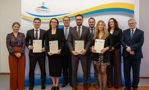 Stojące osoby kobiety i mężczyźni w różnym wieku, niektórzy trzymający teczki papierowe przed sobą 